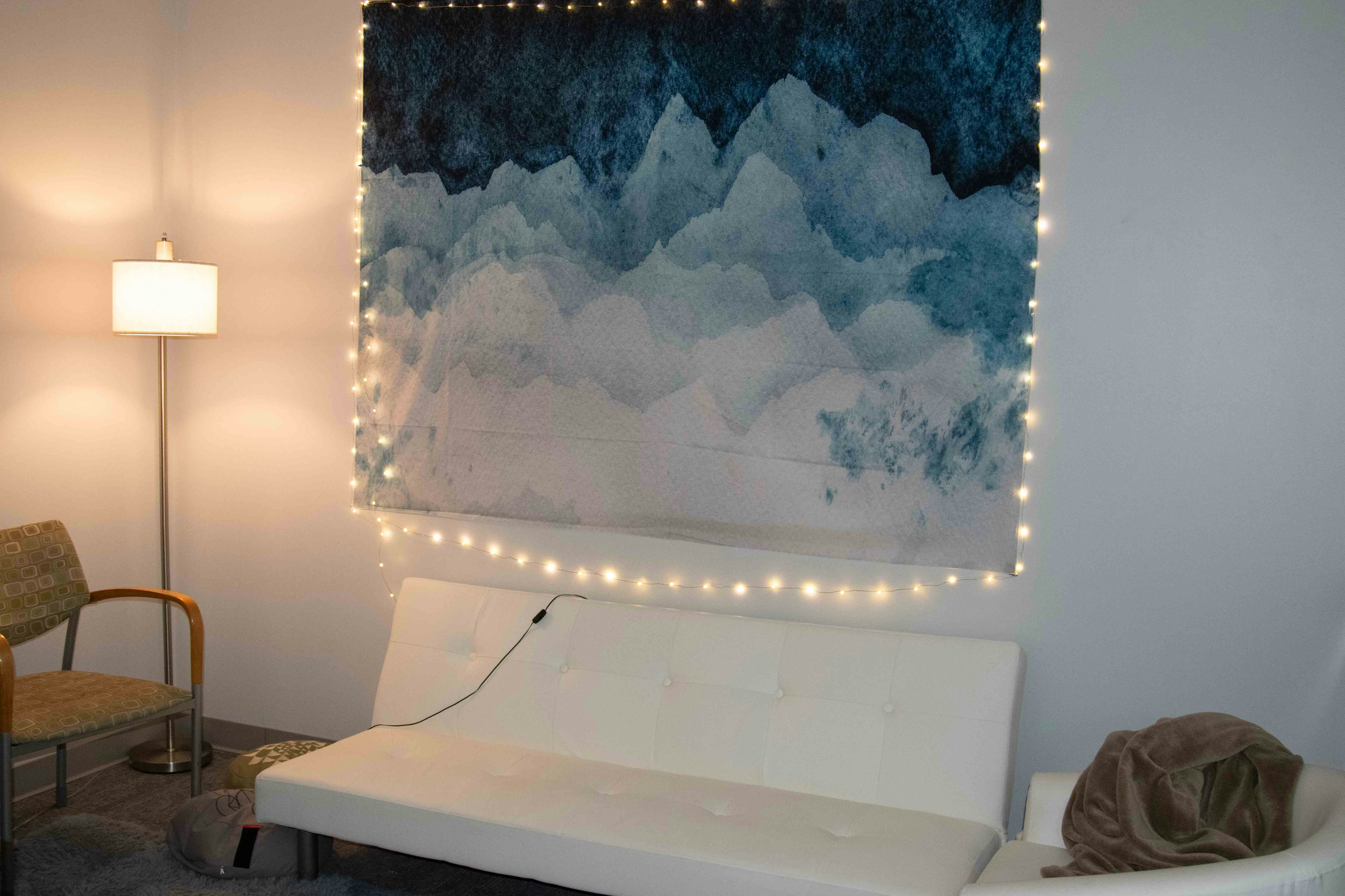Inside of meditation room at Boulder Strong Resource Center. Blue mountain tapestry on wall outlined in twinkle lights. White futon and two chairs.