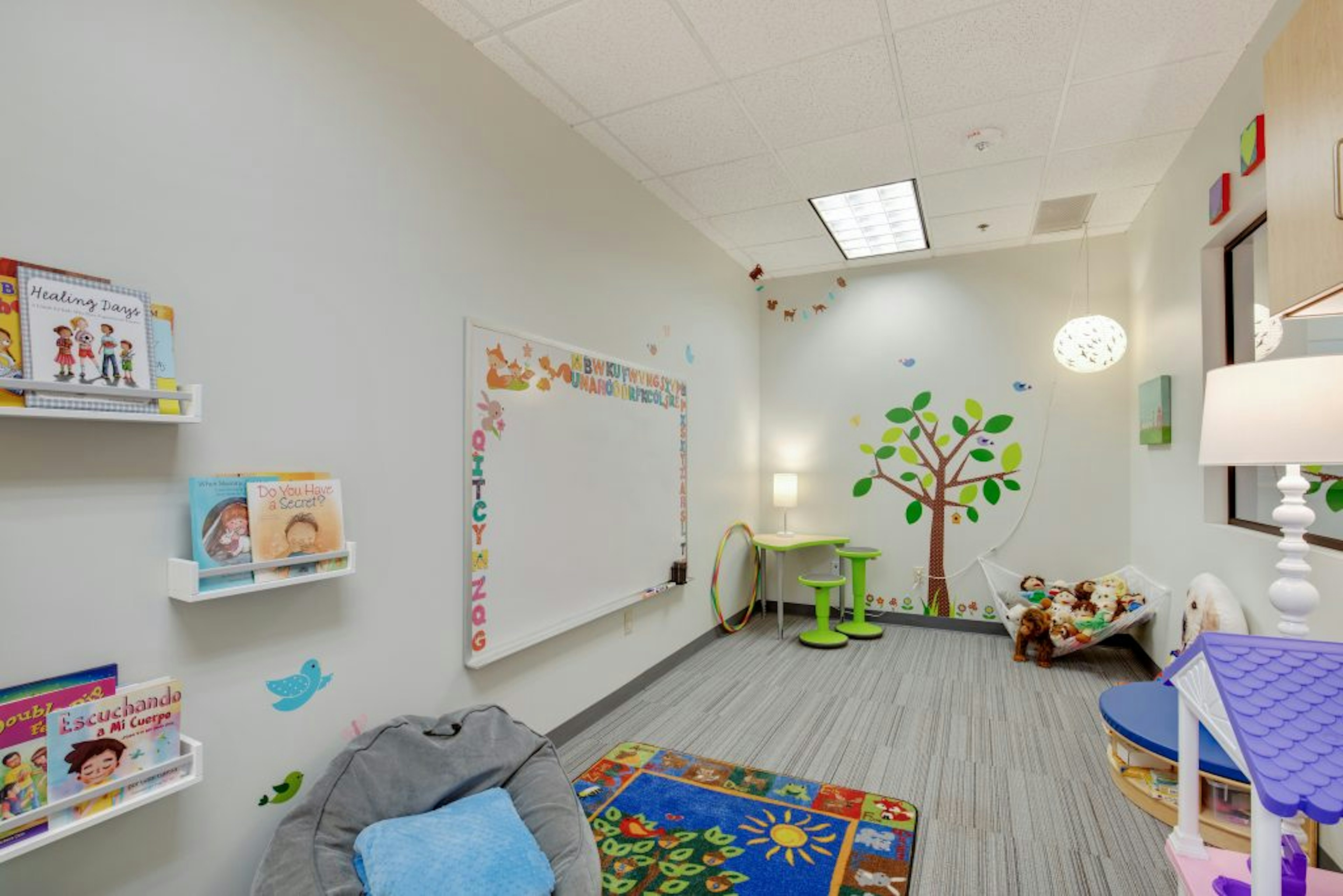 Child's playroom featuring books, colorful rug, hammock full of toys, and mural tree on wall.