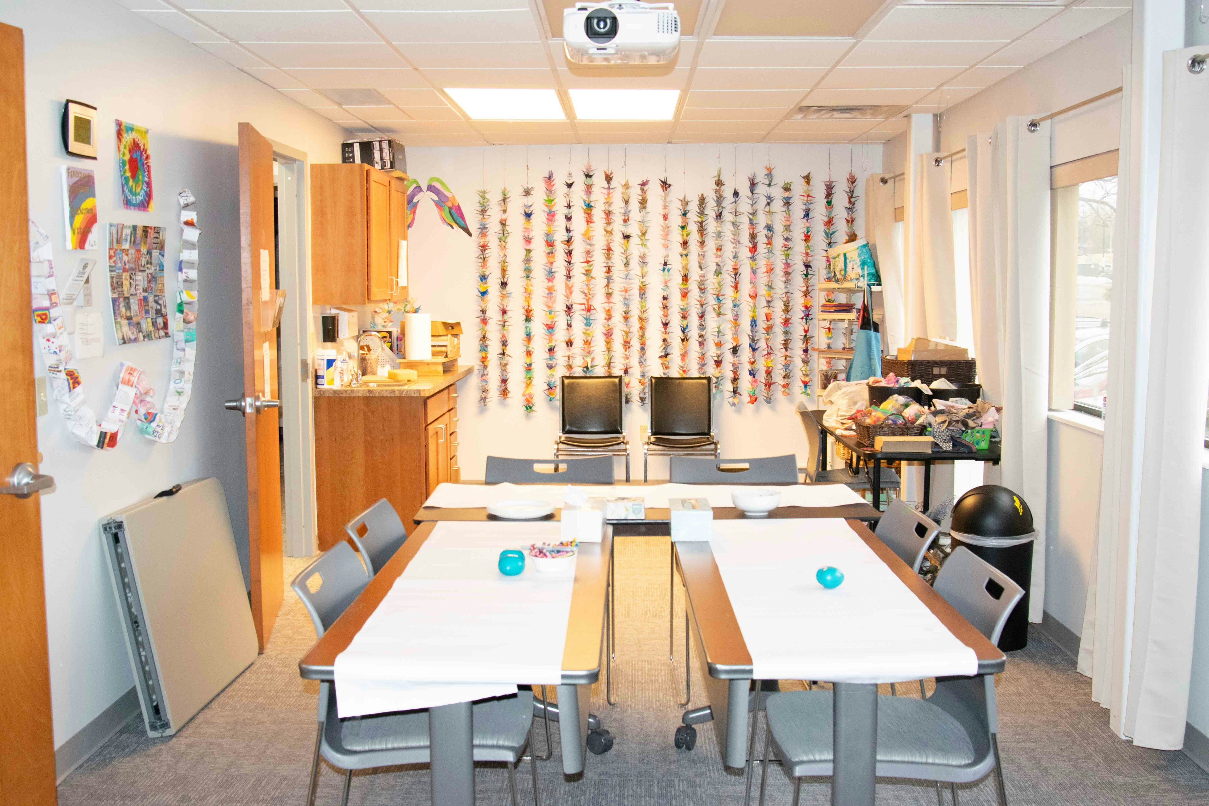 Boulder Strong art room featuring three tables with white protective paper. A wall filled with bird origami, windows with bright white curtains, and a projector hanging from the ceiling.