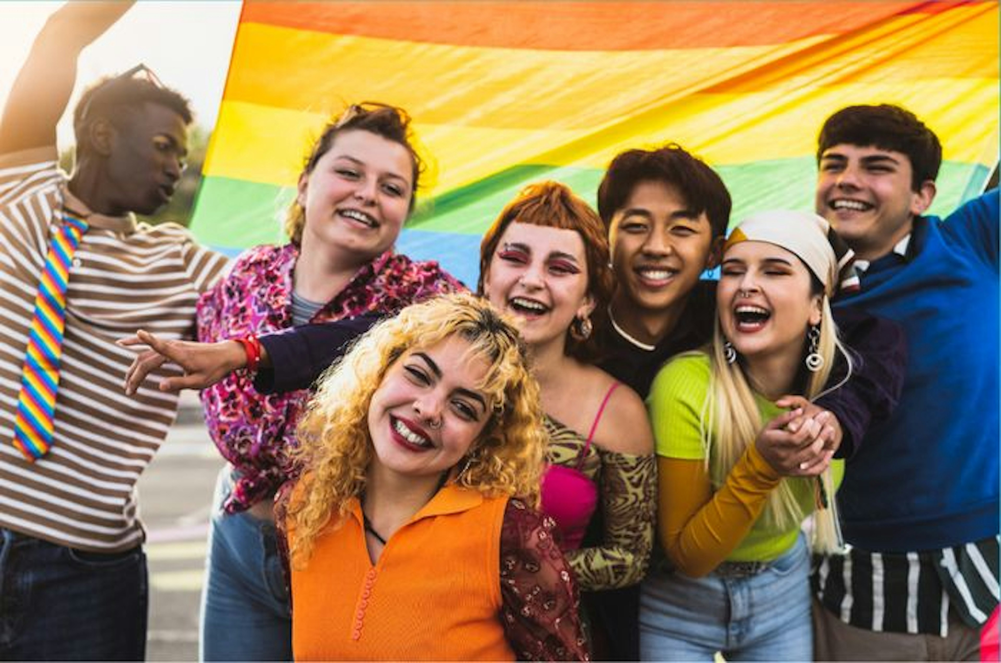 Group of young adults smiling and laughing while holding a pride flag in the background.