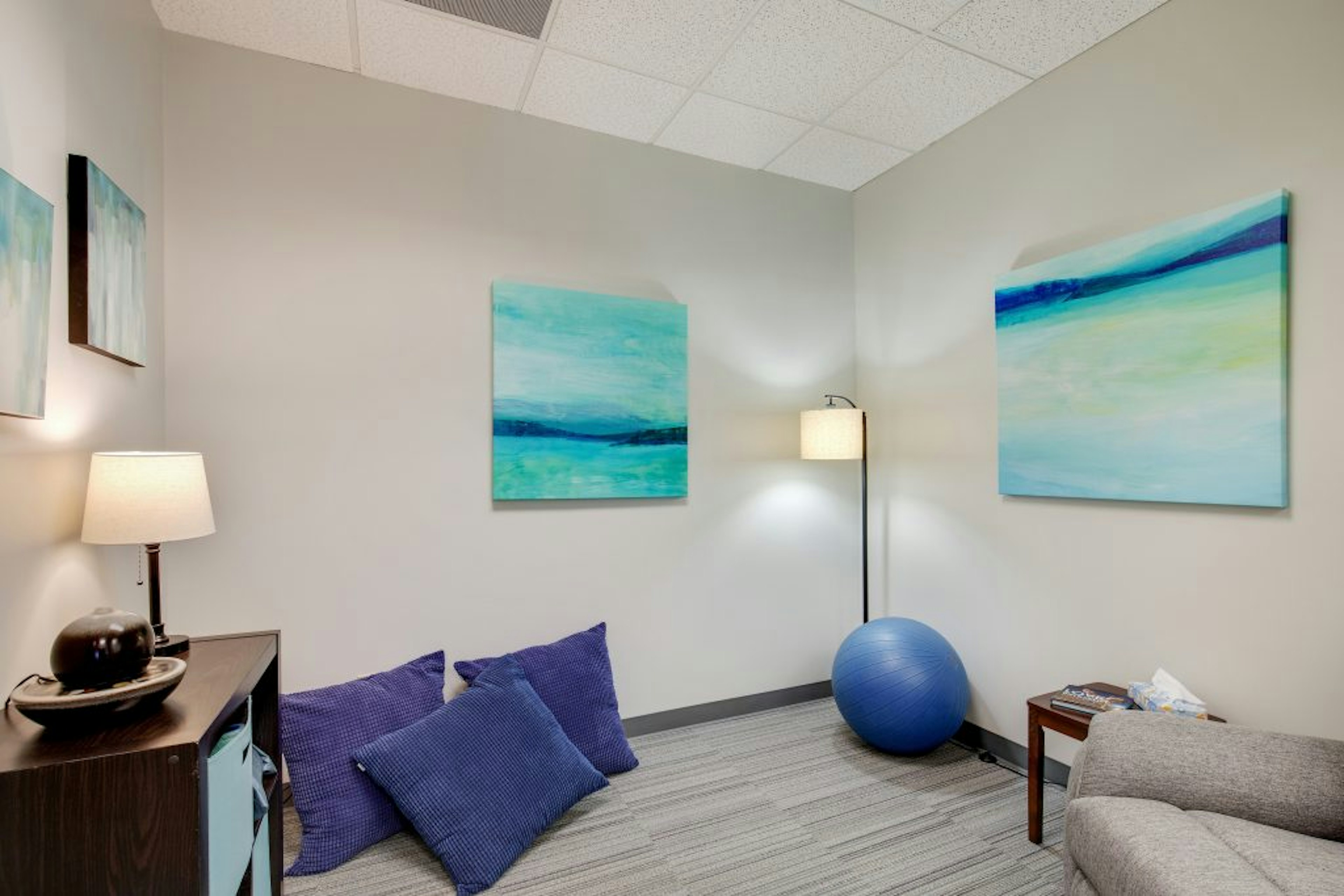 Office with blue pillows leaning up against wall, blue exercise ball, gray couch, and blue art on walls.